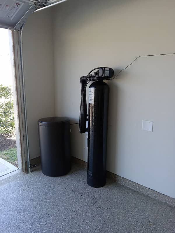 Garage corner with a black water softener tank and a black storage bin beside a wall.