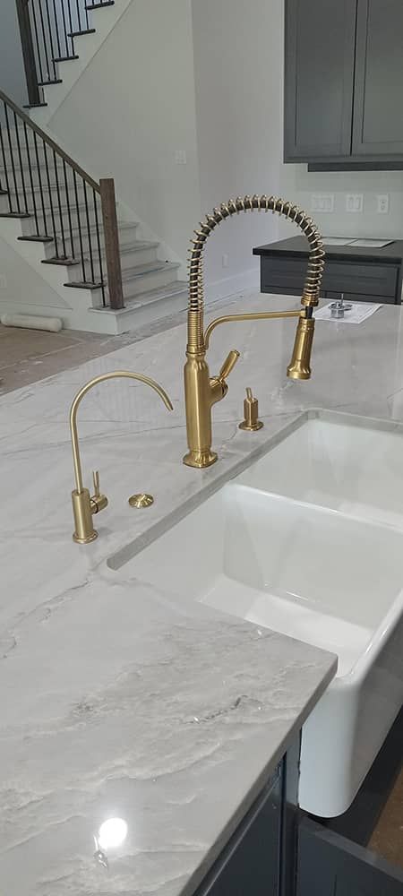 Gold faucet over a white farmhouse sink in a bright kitchen with marble countertop