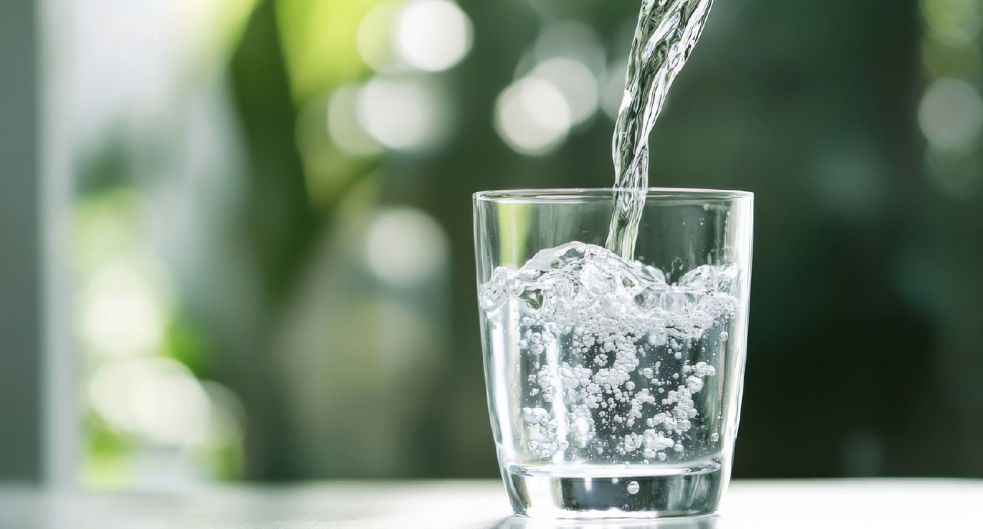 Water being poured into a clear glass on a table, with a blurred green background.