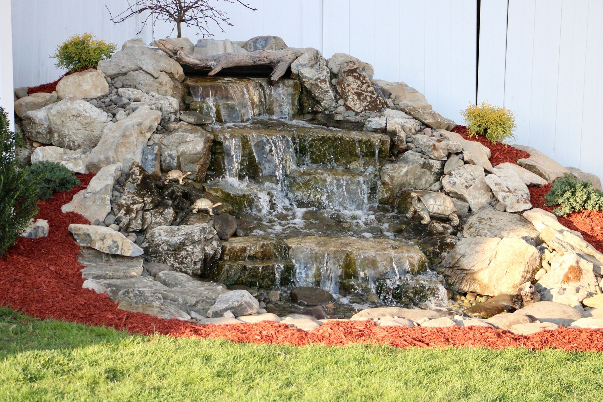 A waterfall is surrounded by rocks and mulch in a backyard.