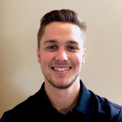 Smiling young man with short brown hair, wearing a black shirt, against a beige wall.