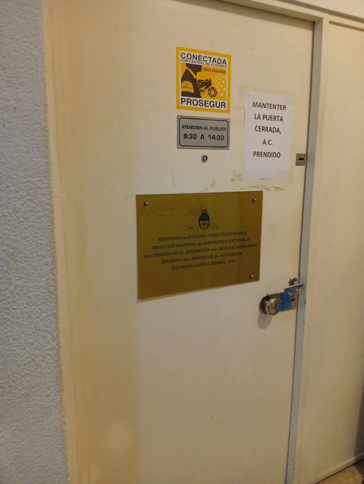 Puerta cerrada de color beige con señales de advertencia y una placa de latón, asegurada con un candado azul en la manija.