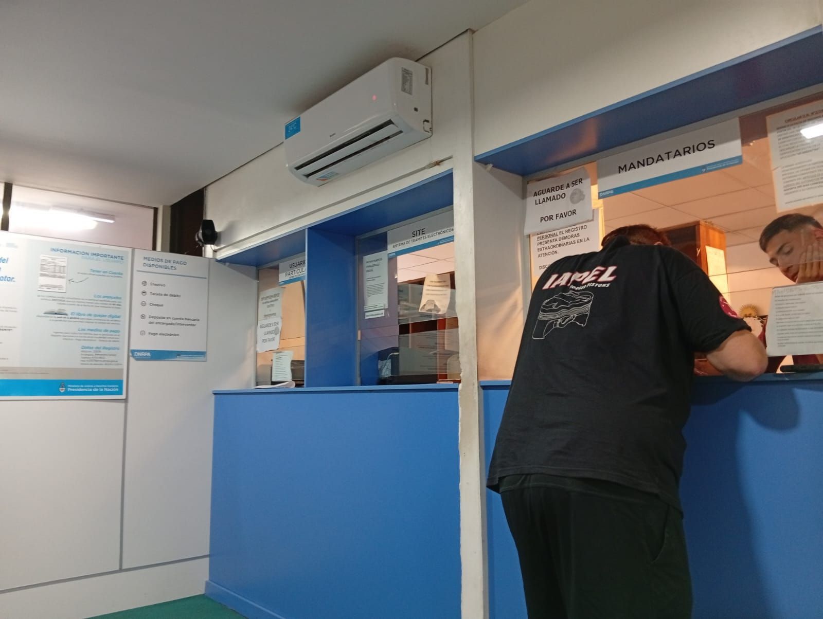 Una persona sentada en un mostrador de servicio azul en una pequeña oficina, hablando a través de una ventana de cristal.