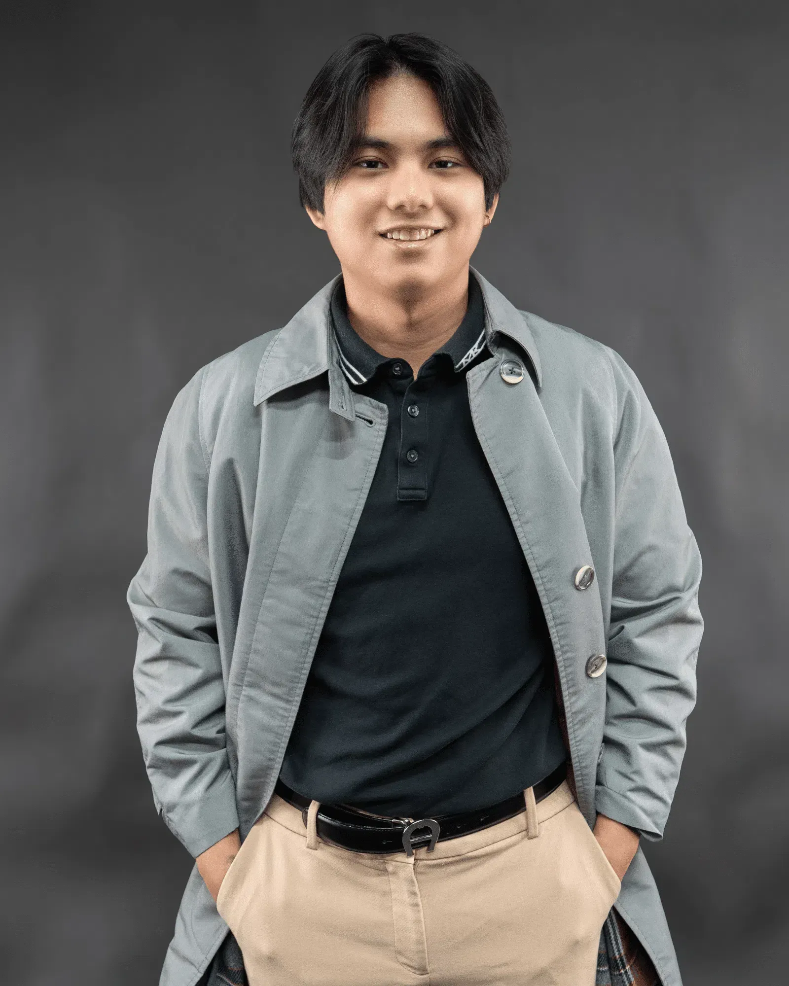 Smiling person in a trench coat over a polo shirt and khaki pants, hands in pockets, against a gray backdrop.
