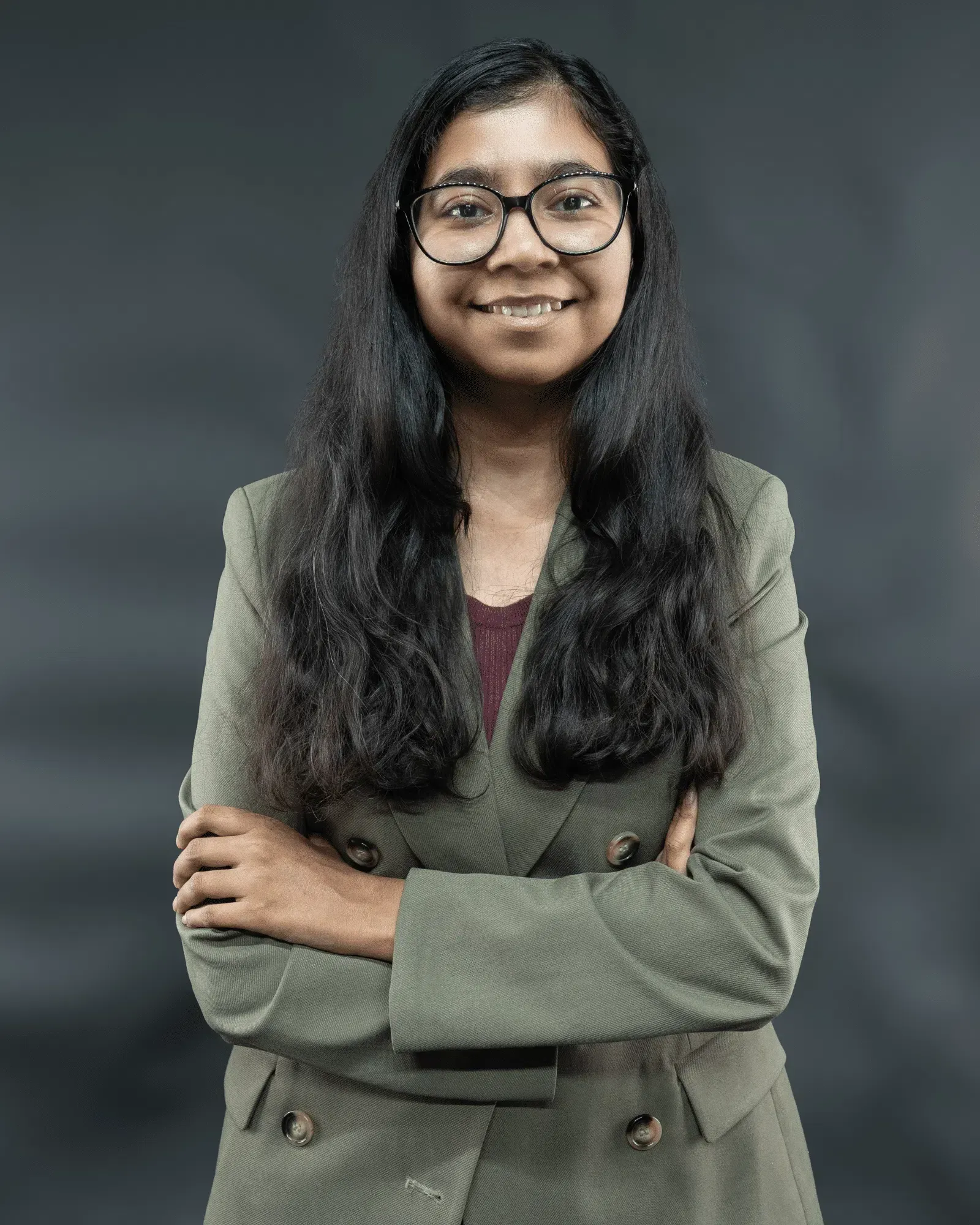 Woman with glasses and arms crossed, wearing a green blazer, smiling, against a gray backdrop.