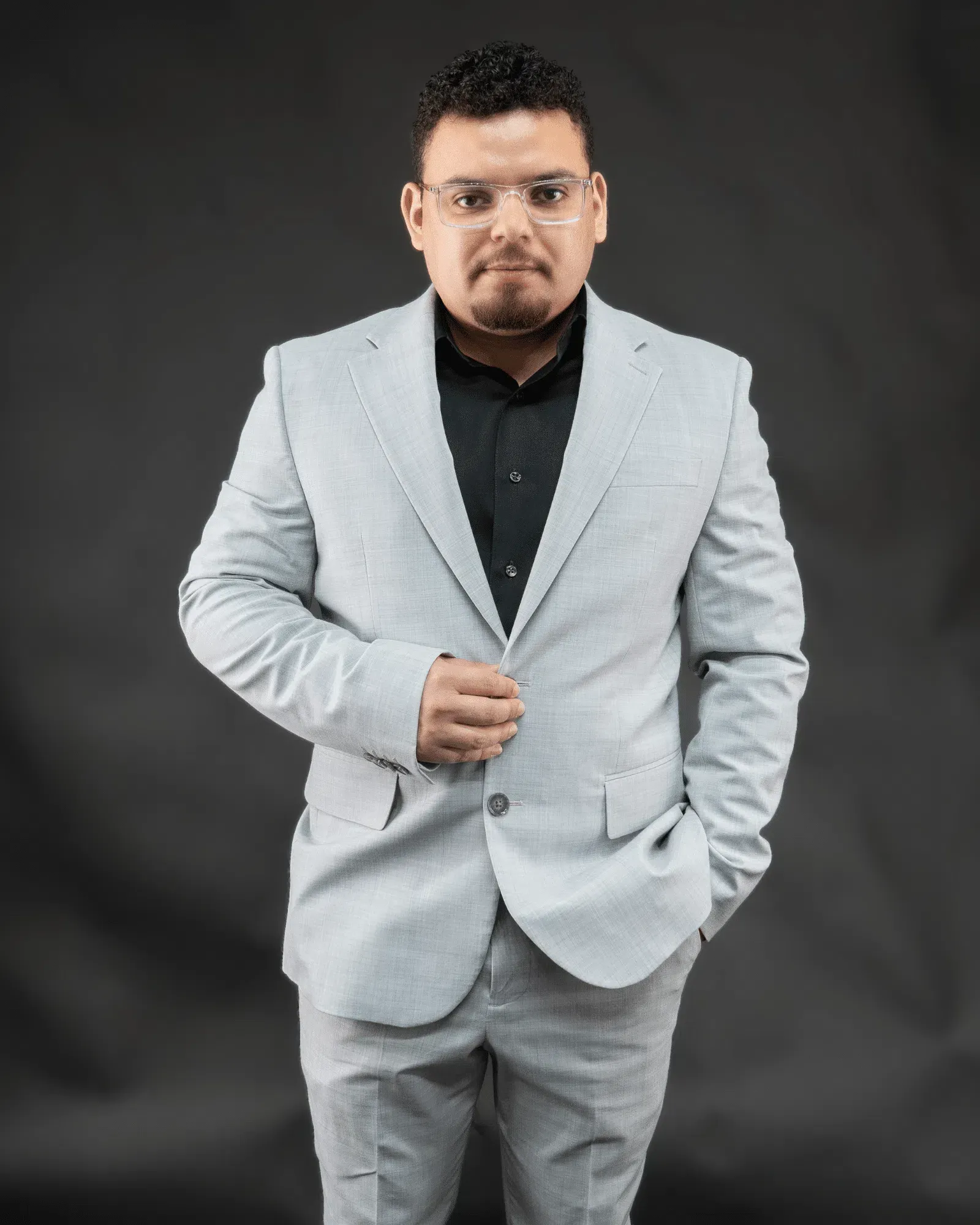 Man in light gray suit and glasses, black shirt, posing against a dark gray background.