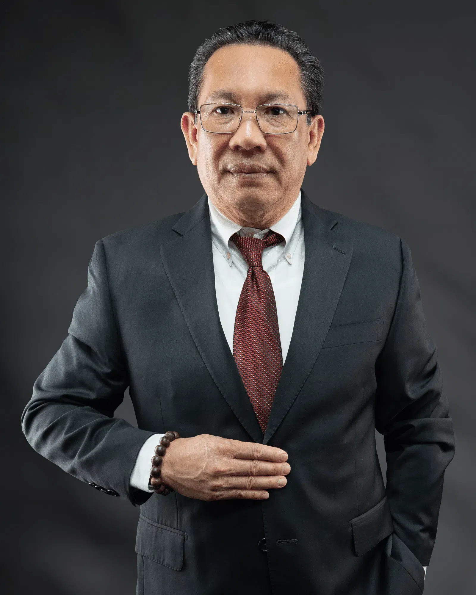 Man in suit, glasses, and tie, hand on chest, posing against a dark background.