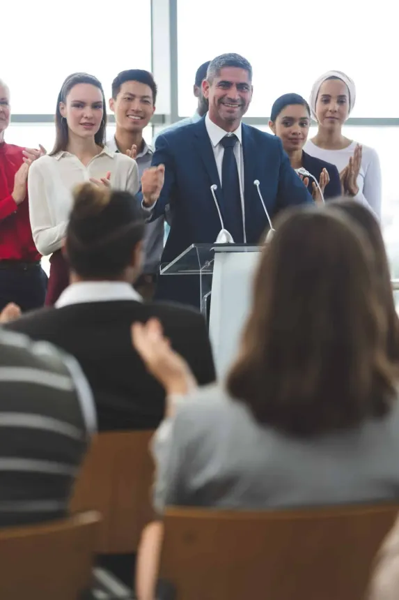 Man in suit speaking at podium, surrounded by people clapping in an indoor setting.
