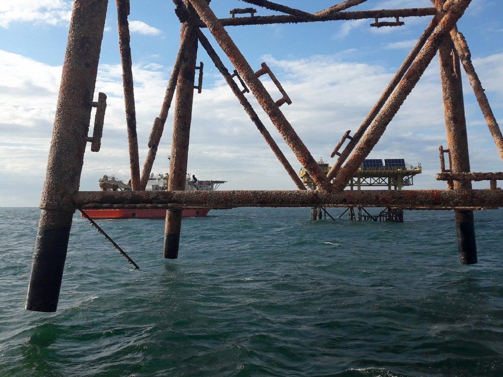 Rusty offshore oil platform legs in ocean with a ship and other platforms in the distance.