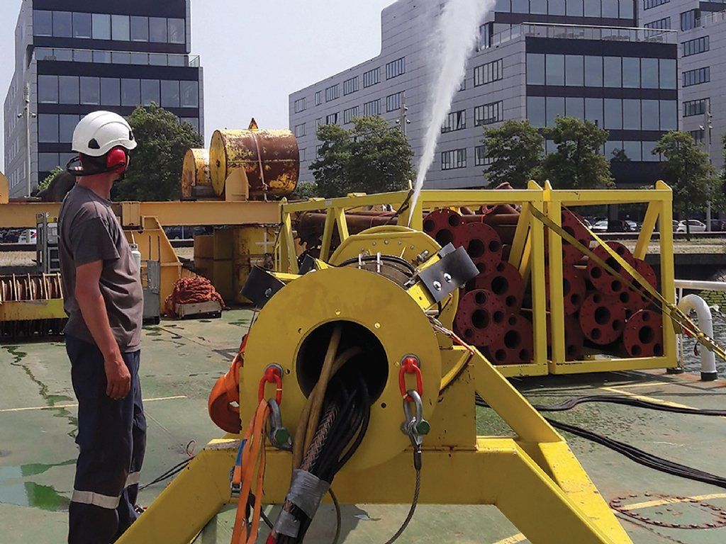 Man observing high-pressure water test from yellow machinery on a platform. Buildings in background, sunny day.