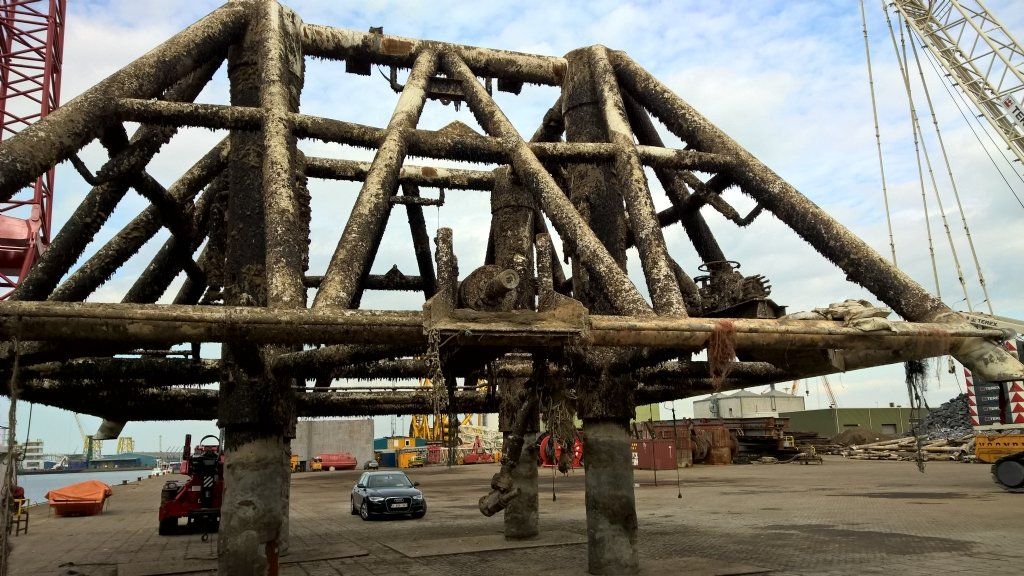 A barnacle-covered, geometric steel structure on concrete supports, possibly a pier component, in a port.