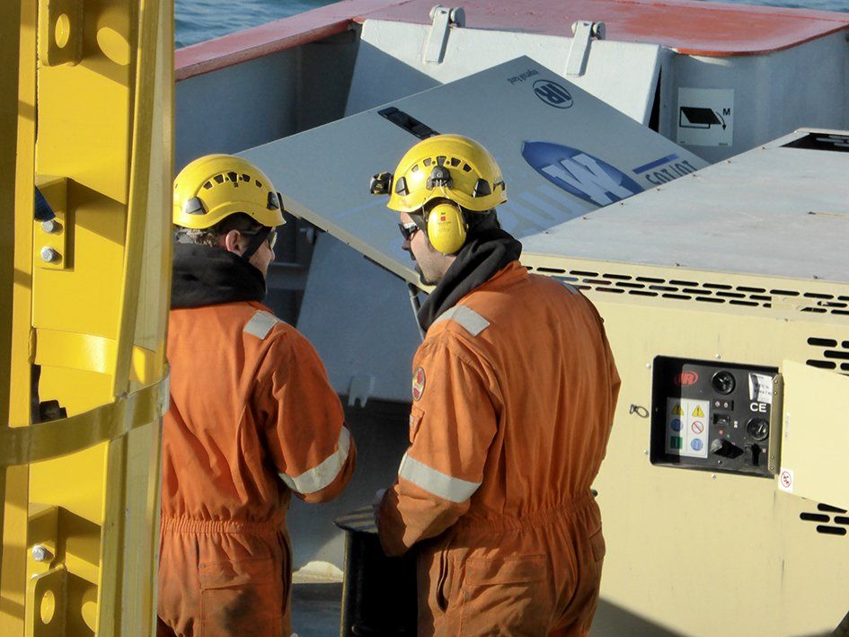 Two workers in orange jumpsuits and yellow helmets on a ship deck, near a generator.