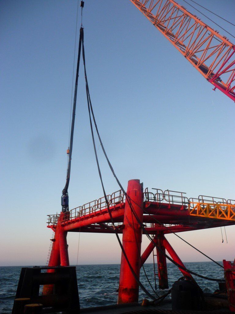 Red offshore structure being lifted by a crane against a blue sky, water below.