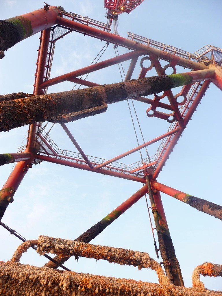 Red offshore oil rig against a blue sky, covered in barnacles and rust.
