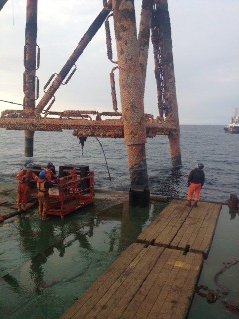 Offshore oil rig platform with workers in orange suits. Water surrounds the rusty structure. A wooden walkway extends.