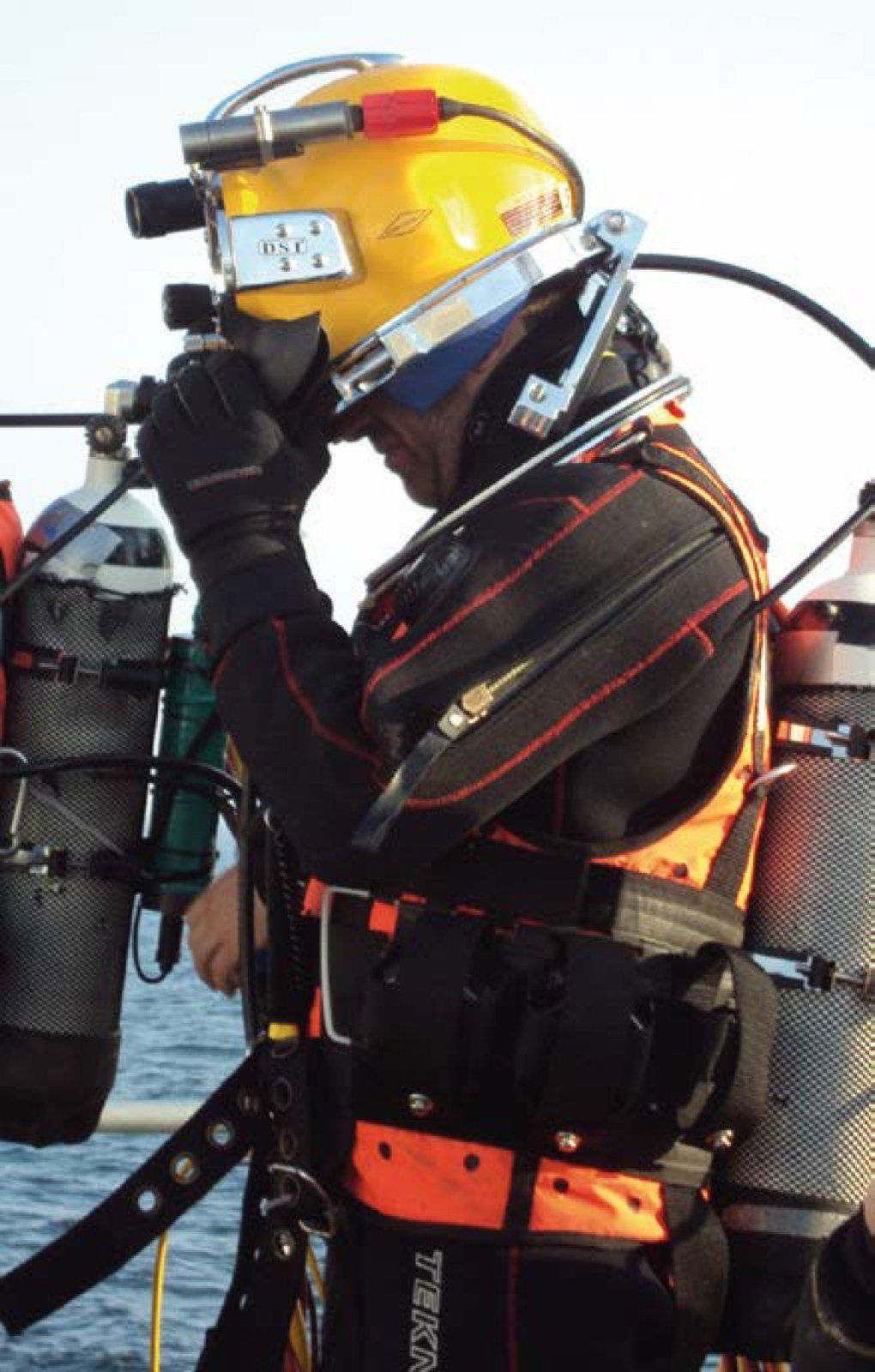 Diver in yellow helmet and black suit, on a boat, adjusting gear with tanks on back.