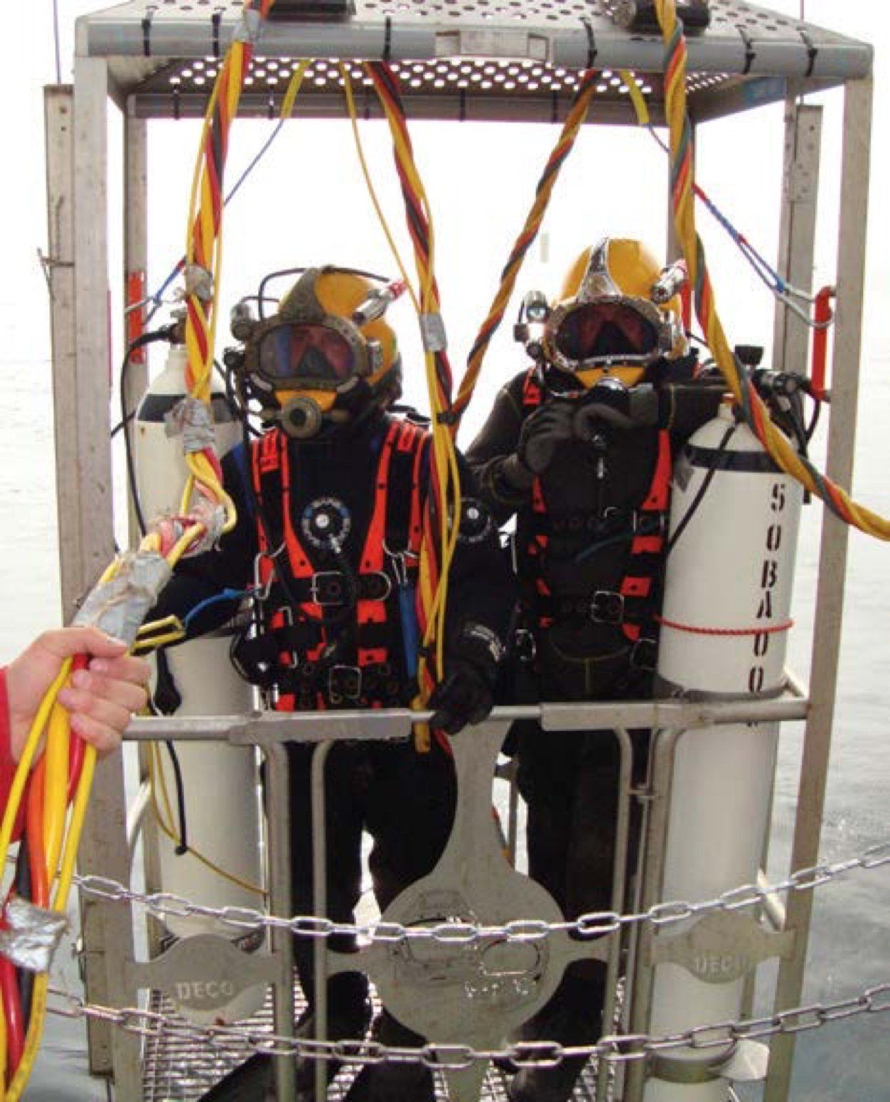Two divers in cage, wearing helmets and diving suits, prepared to descend into water.