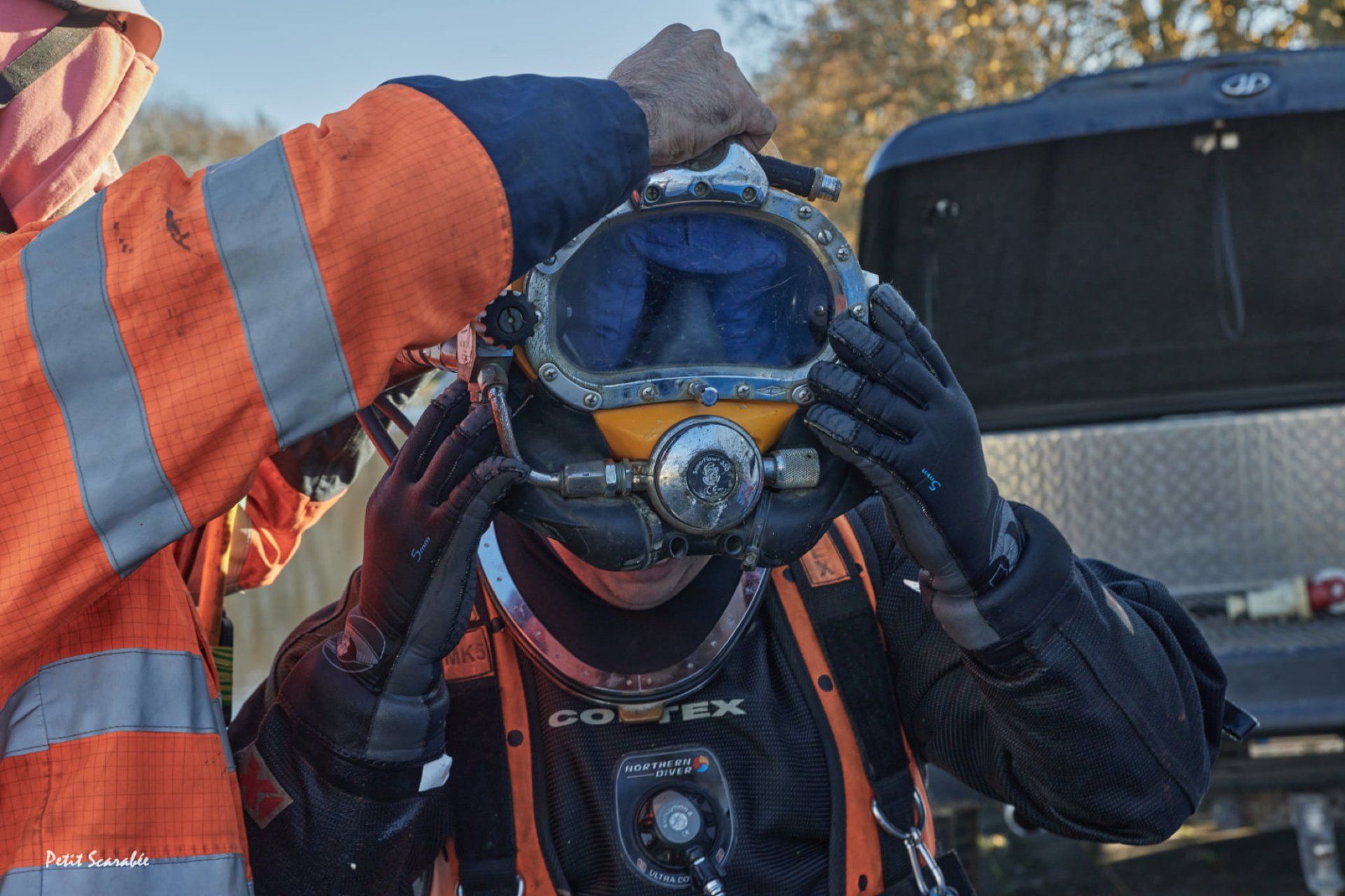 Diver being helped to put on a diving helmet, orange suit, blue visor, outdoors.