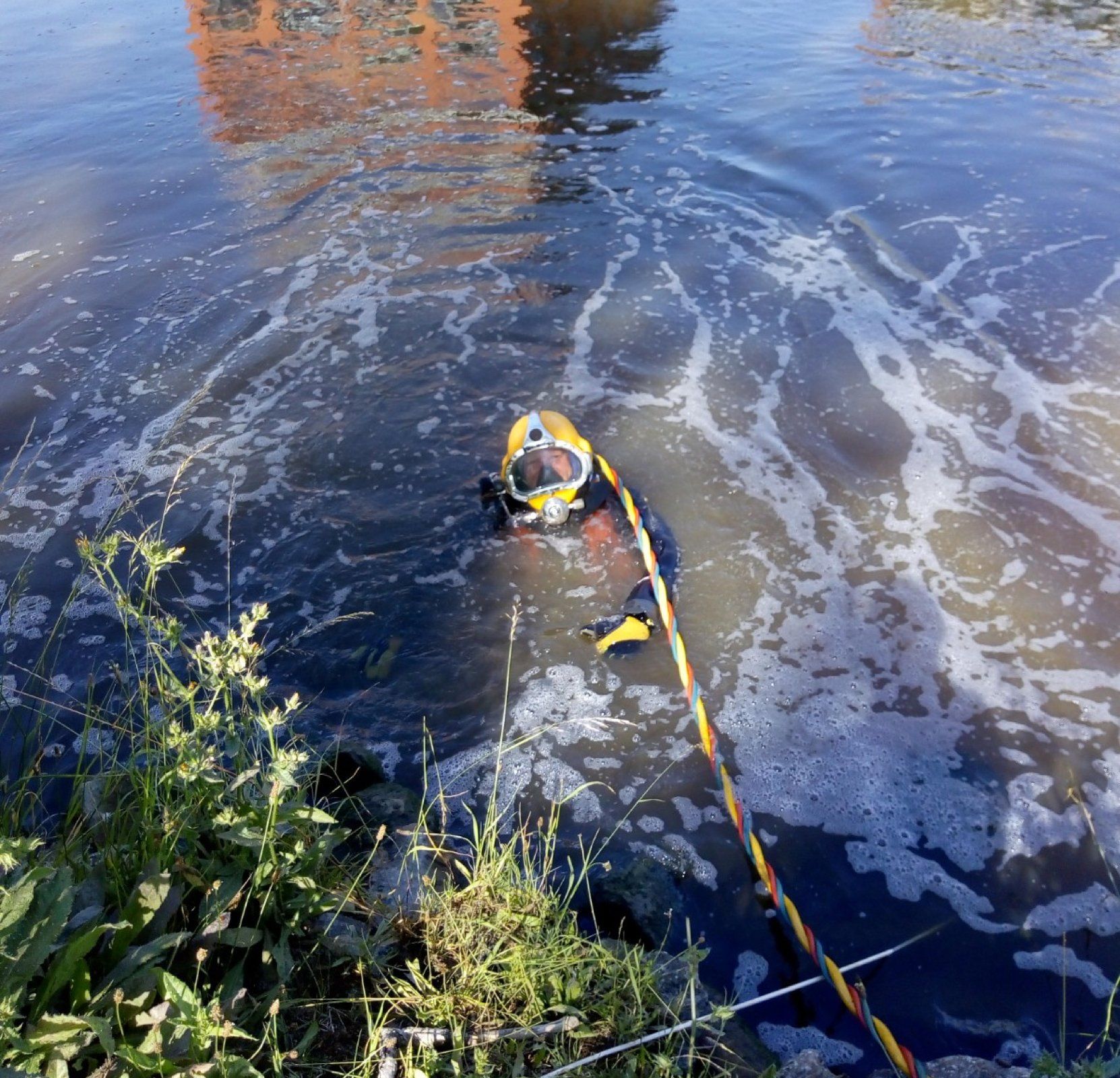 Diver in dark water, wearing yellow helmet, connected by rope. Vegetation in foreground, building reflected in the water.