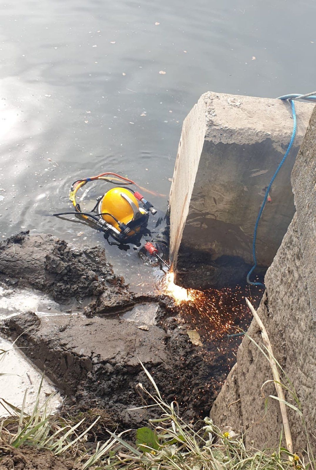 Diver in yellow helmet welding underwater on a concrete structure, sparks visible, near dark water and shoreline.