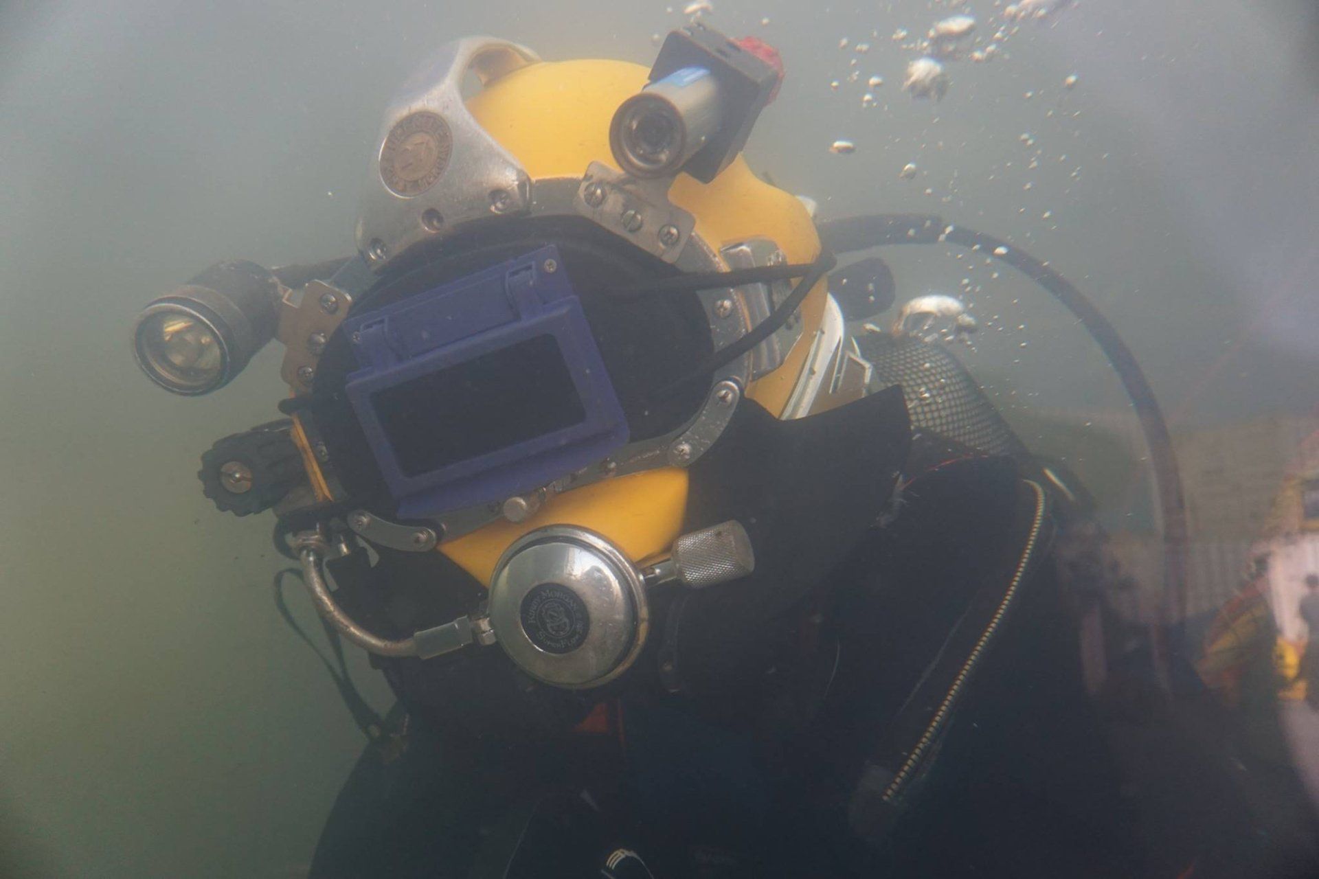 Diver in a yellow helmet, underwater, with lights and a screen. Water is murky.