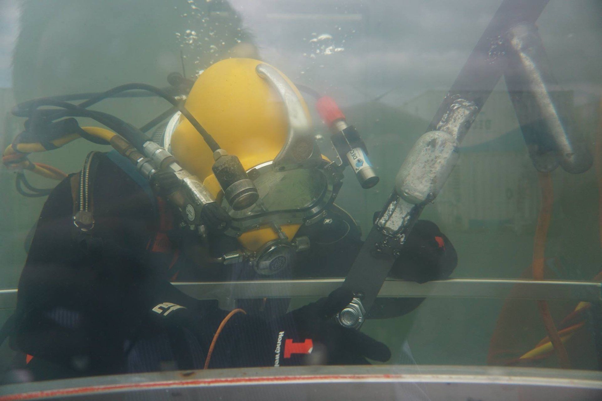Diver in yellow helmet and black suit inside a clear enclosure, holding a metal bar underwater.