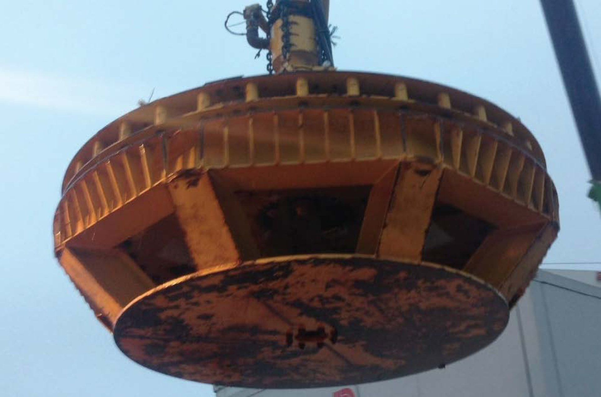 Large, round, yellow metal electromagnet suspended by a crane against a light blue sky.