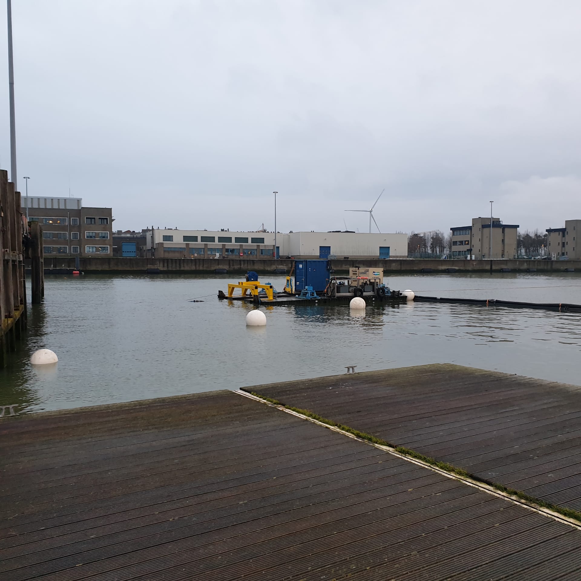 Water with industrial equipment, floating buoys, and buildings under cloudy sky.