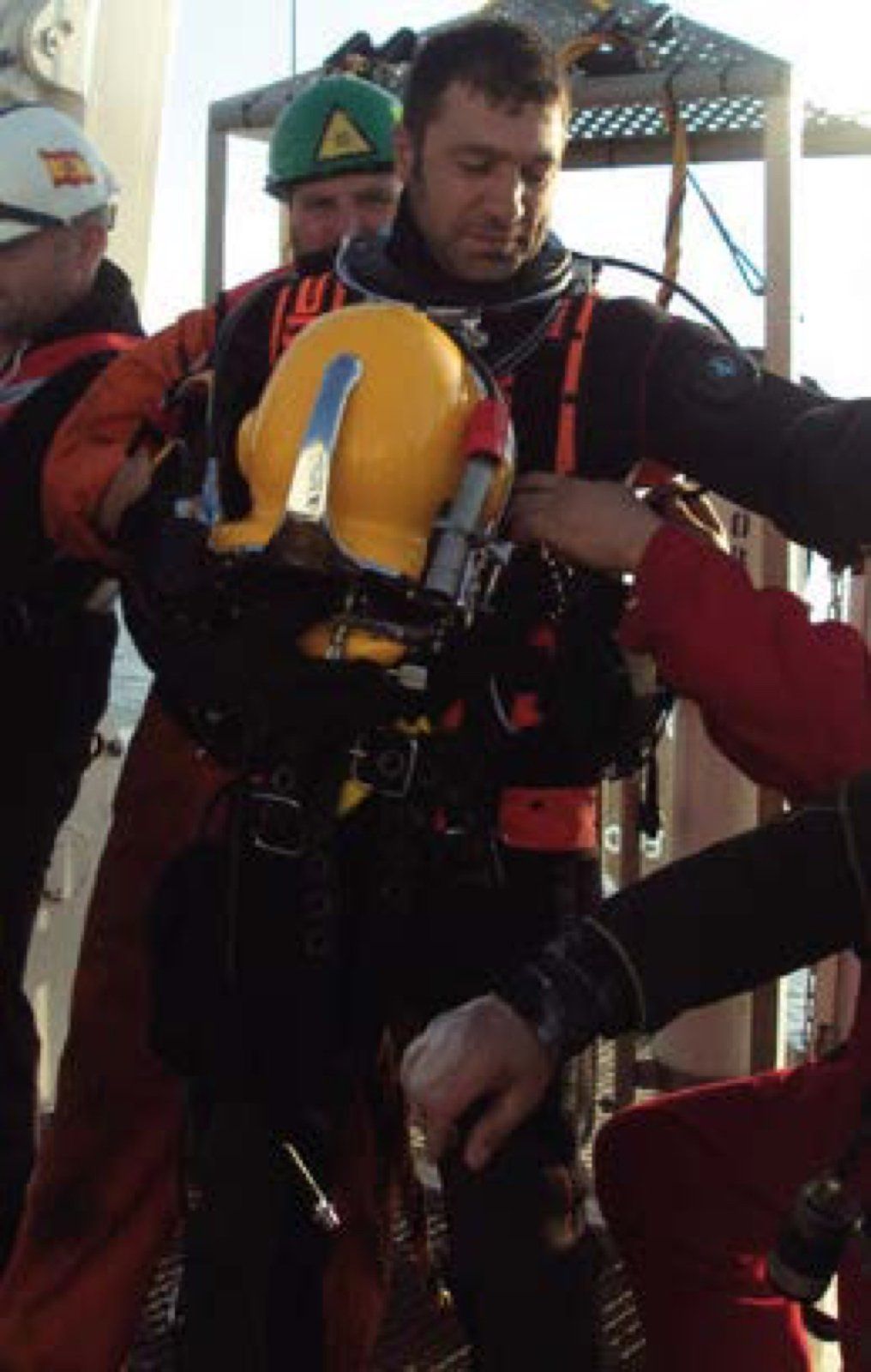 Person in diving suit, being assisted by others, wearing a yellow helmet, on a platform.