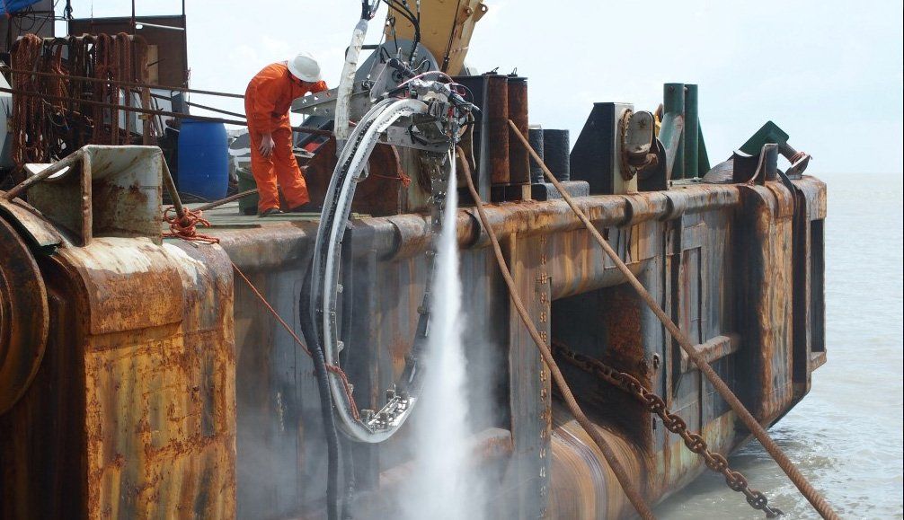 Person in orange suit operating water cutter on rusty barge near water.