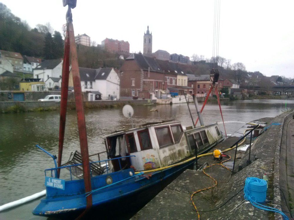 A blue and white boat being lifted from a river by a crane, buildings in the background.