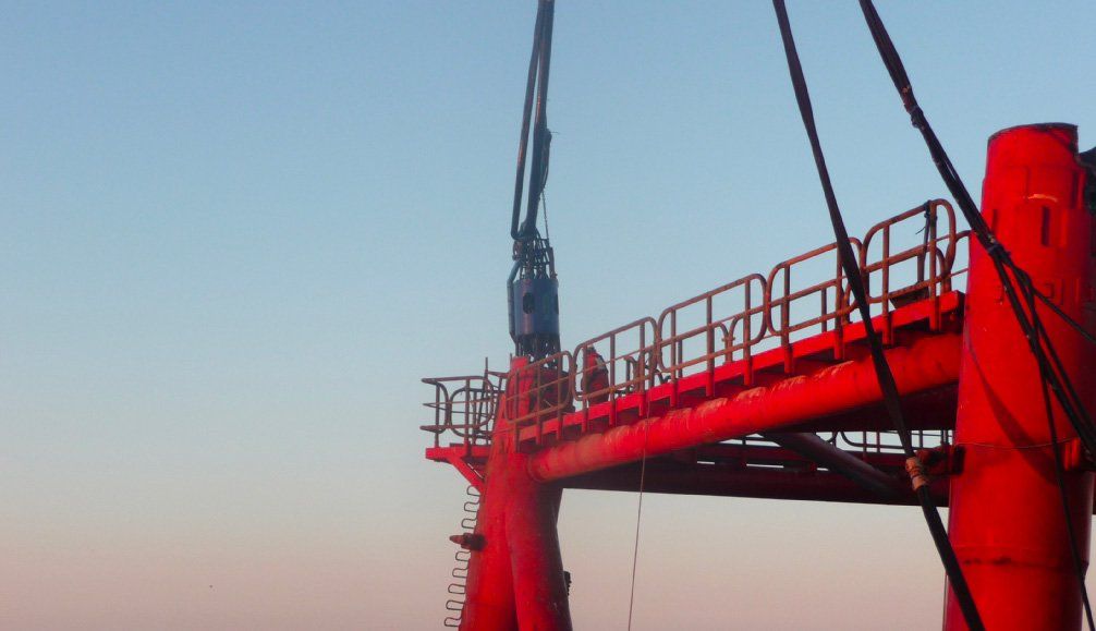 Red offshore oil platform with crane lowering equipment against a pale blue sky.