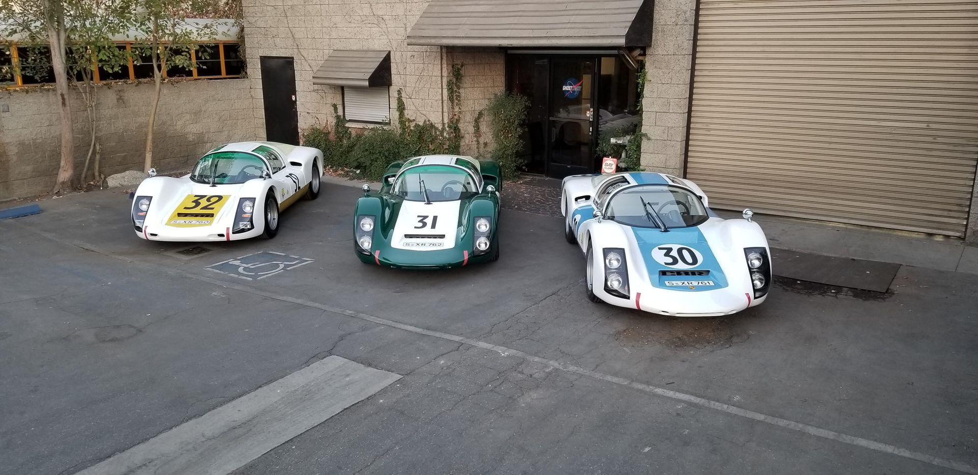 Ford v Ferrari cars - Three vintage race cars parked outside a building. One white with yellow, one green, one white with blue.