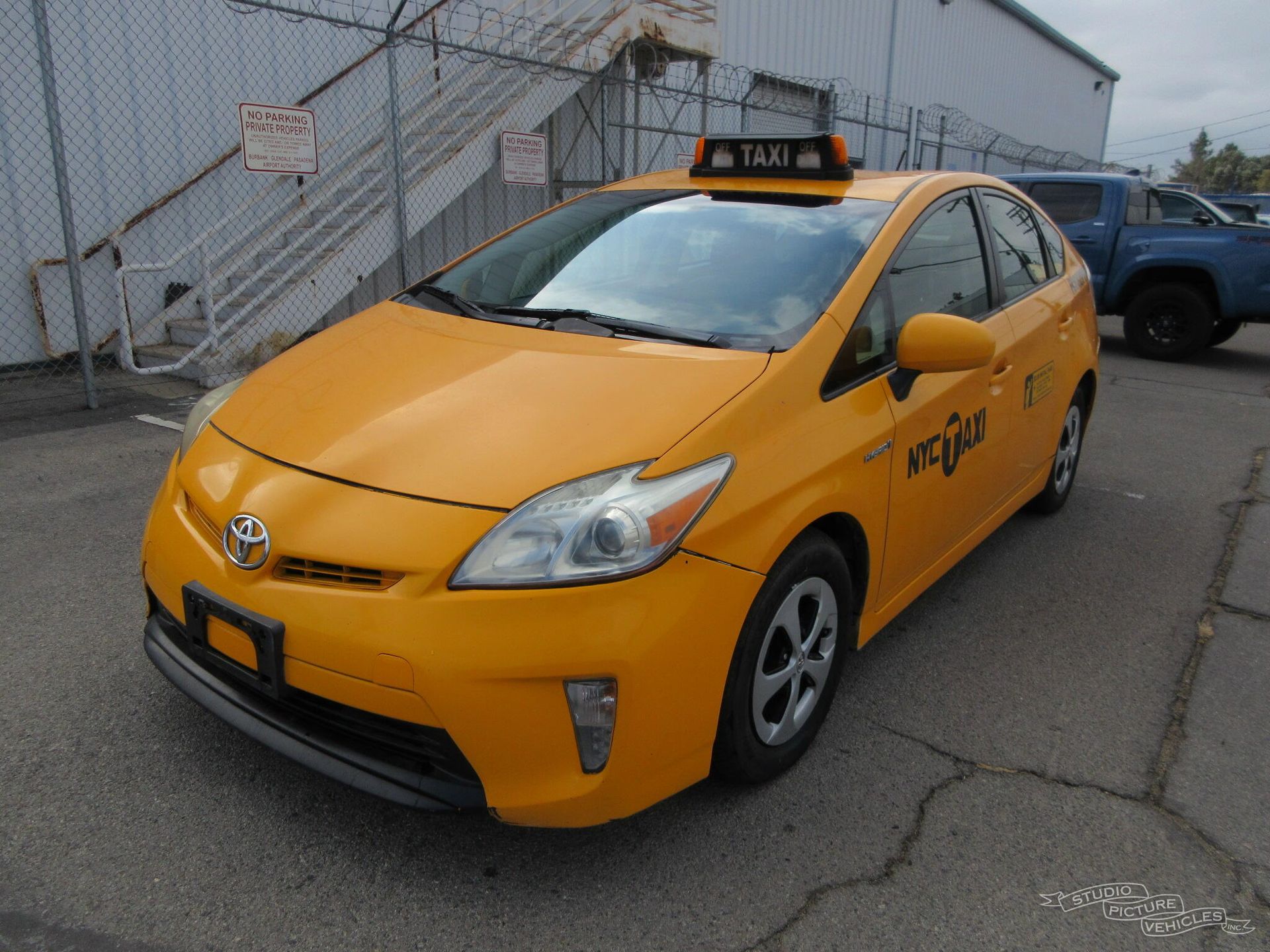 A yellow taxi cab is parked in front of a chain link fence and ready to install a pod rig for film shooting.