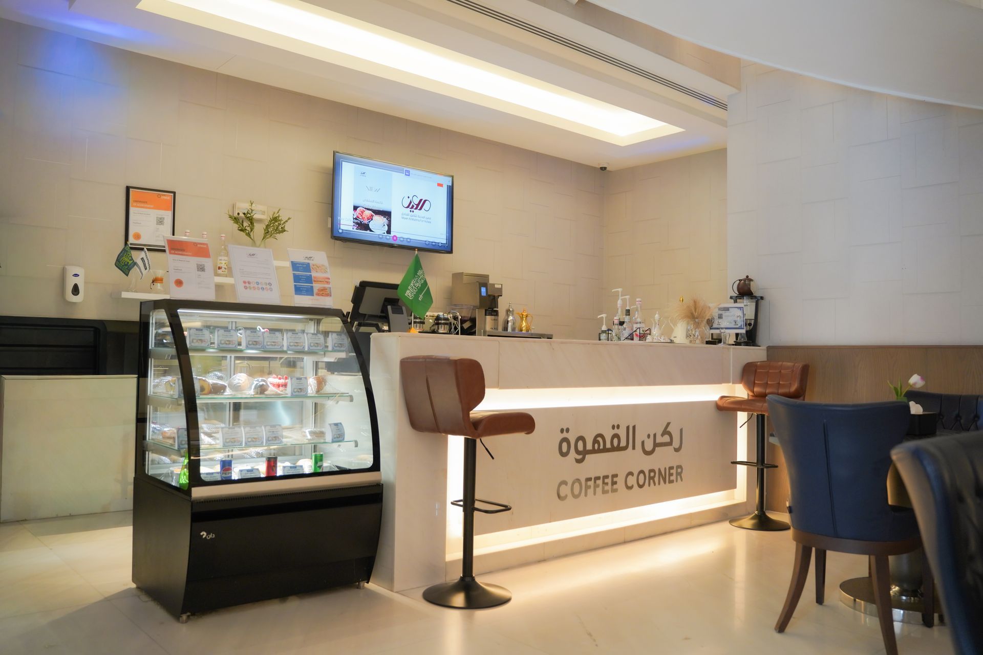Modern café counter with display case, lit sign, menu screens, and seating in a bright interior.