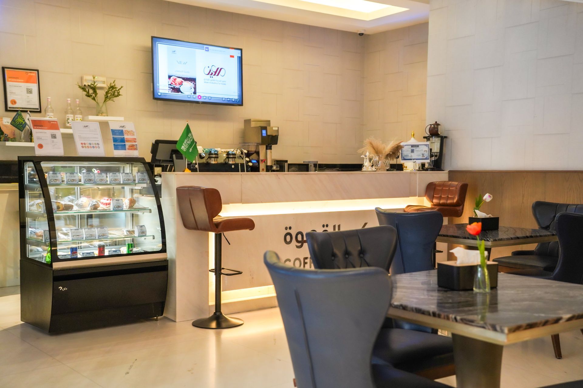 Modern café interior with a counter, display fridge, wall menu screen, and seating area