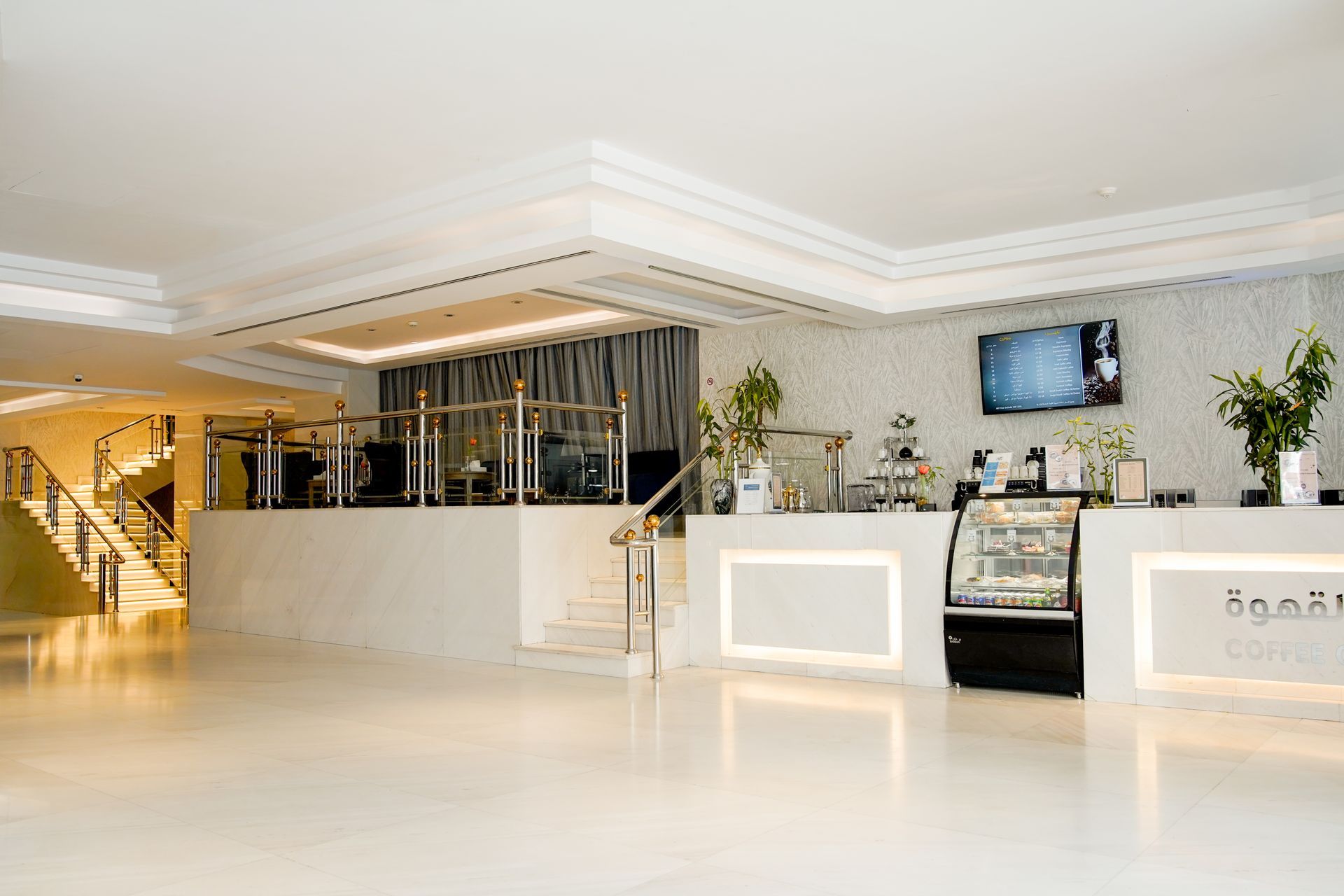 Bright modern hotel lobby with front desk, staircase, seating, and TV screens