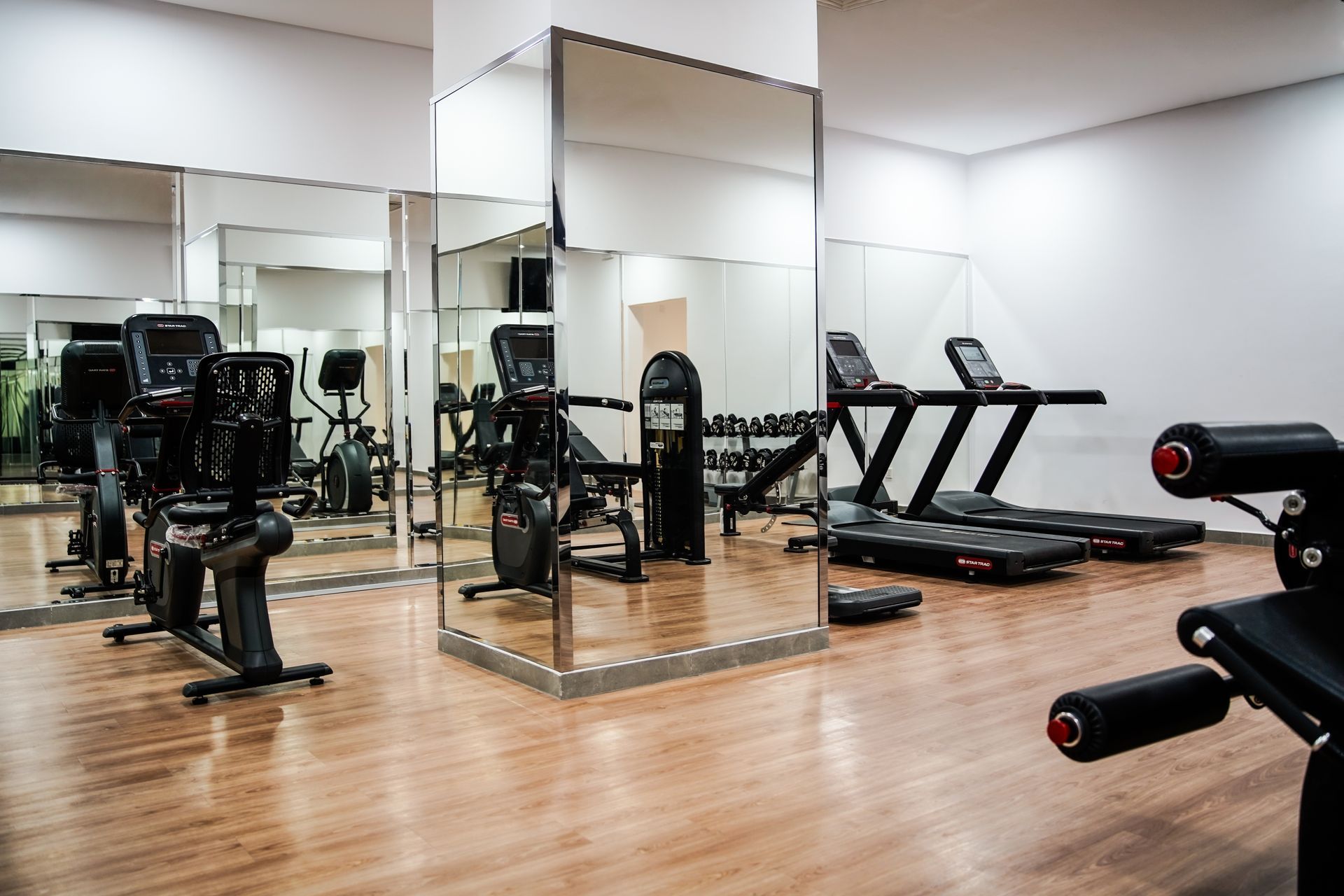 Empty gym room with mirrored walls, treadmills, and weight machines on a wooden floor.