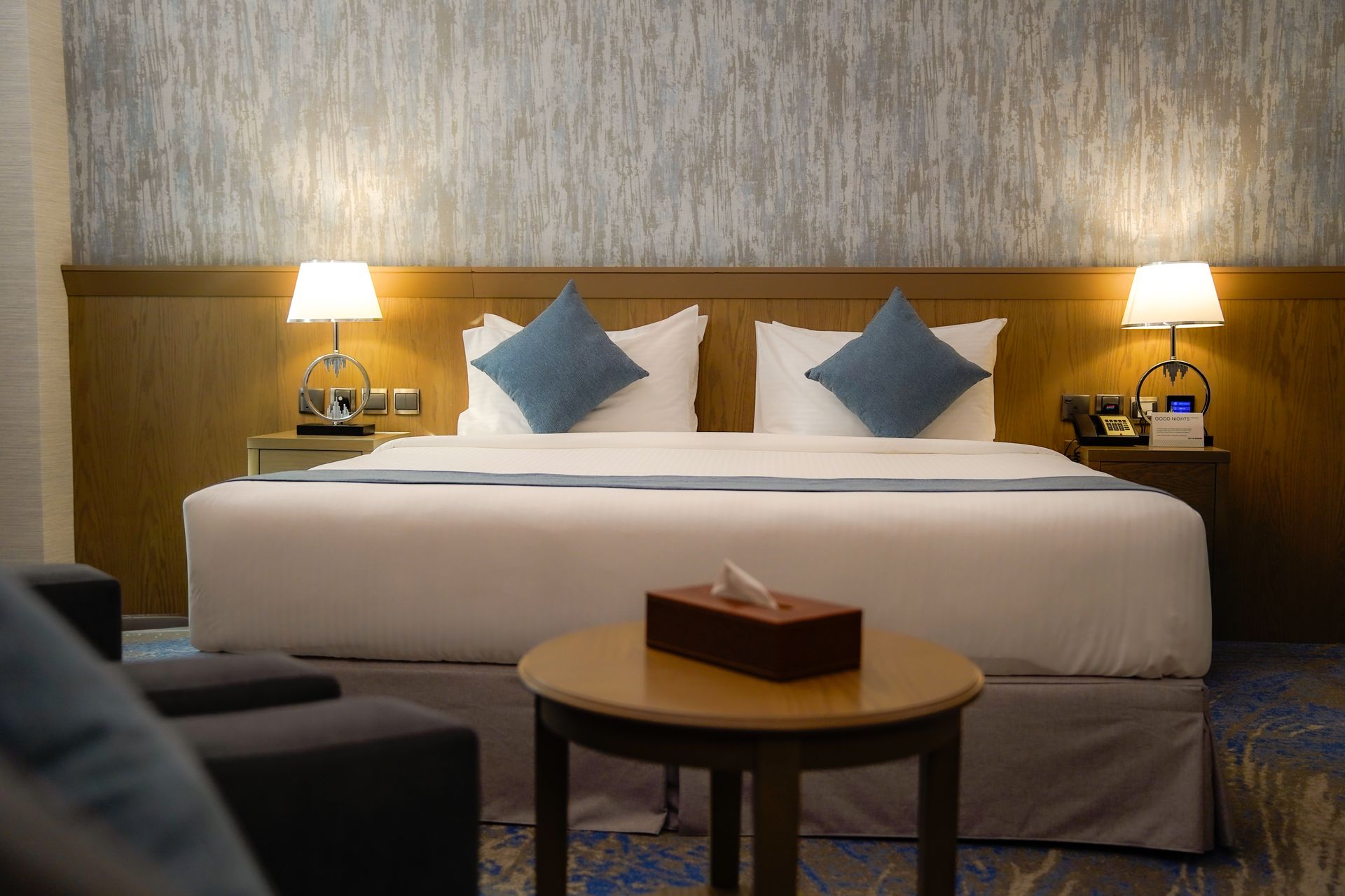 Modern hotel room with a neatly made bed, blue pillows, bedside lamps, and a small round table.