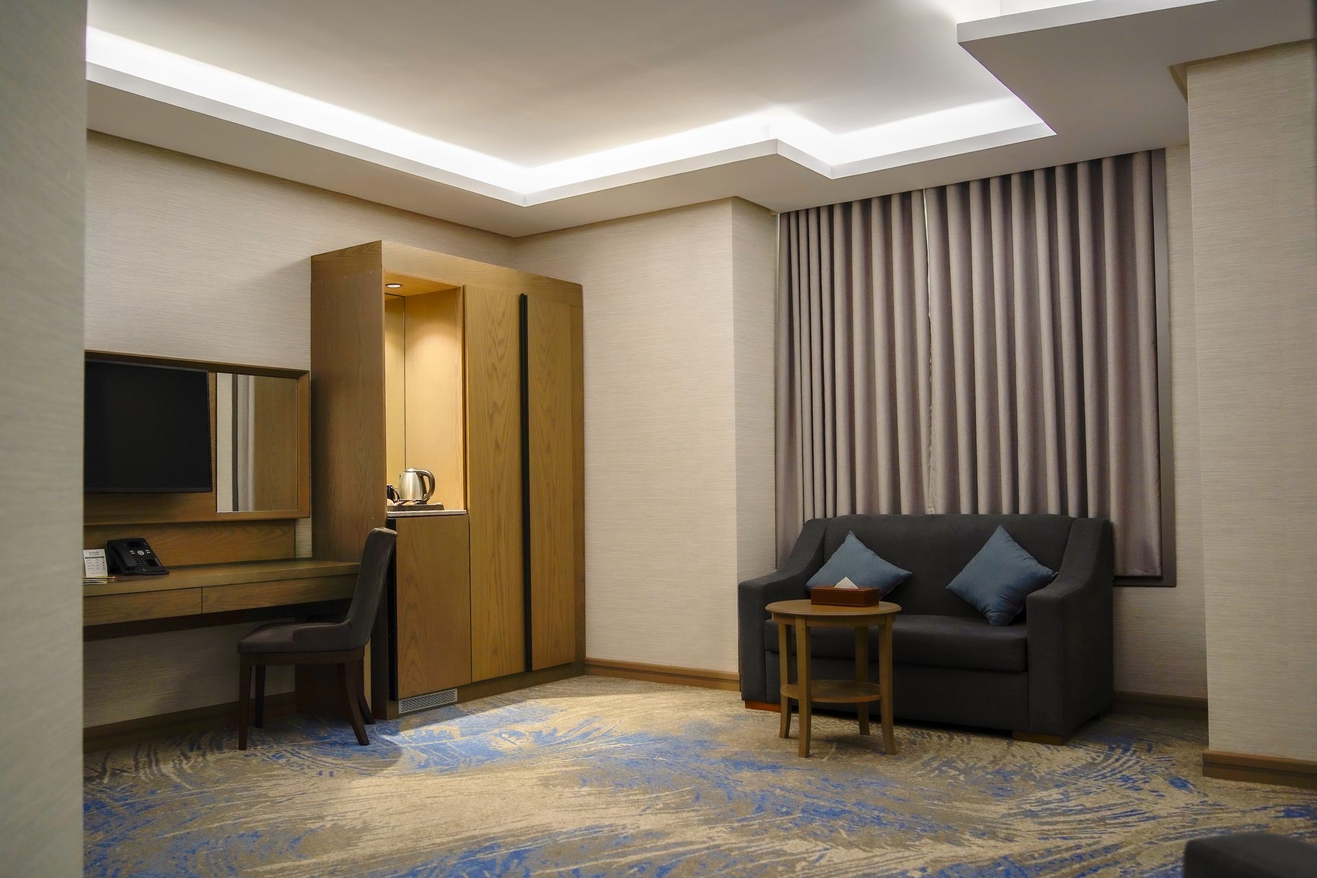 Modern hotel room with desk, sofa, curtains, and soft ceiling lighting