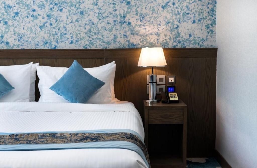 Neatly made hotel bed with blue pillows, floral wallpaper, and a lit bedside lamp.