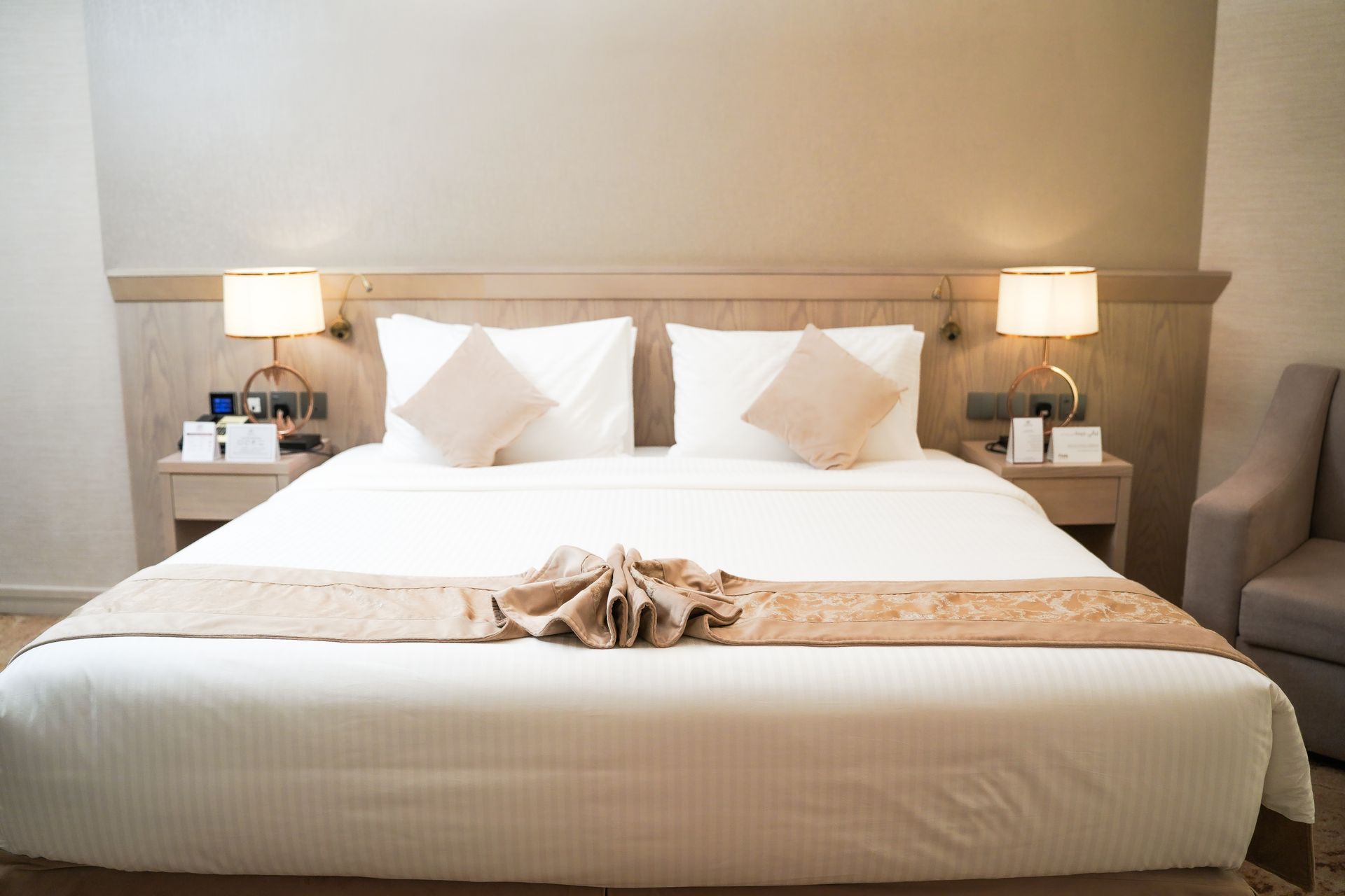 Neatly made hotel bed with white linens, beige accents, bedside lamps, and a folded towel centerpiece
