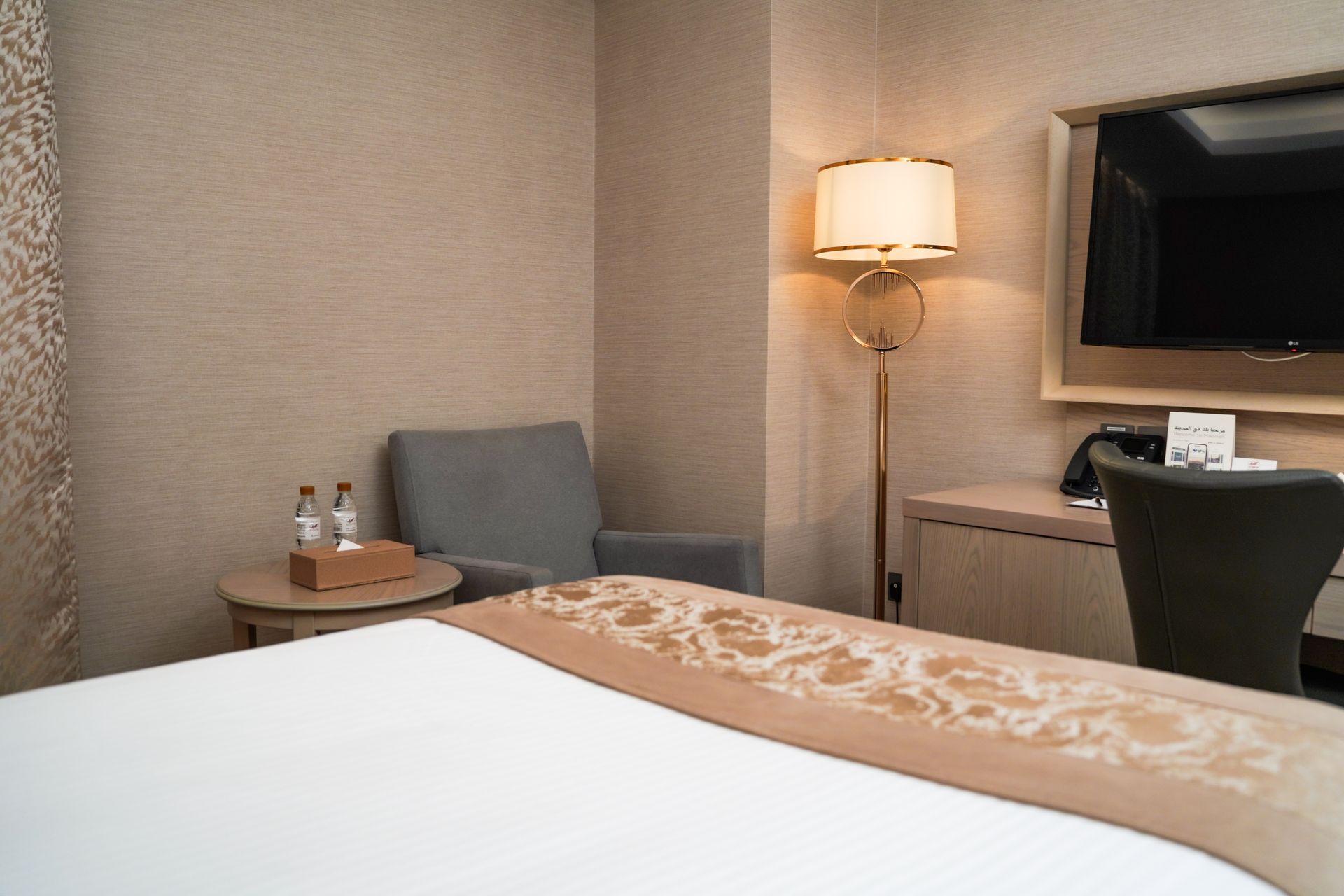 Modern hotel room with a bed, armchair, lamp, desk, and wall-mounted TV