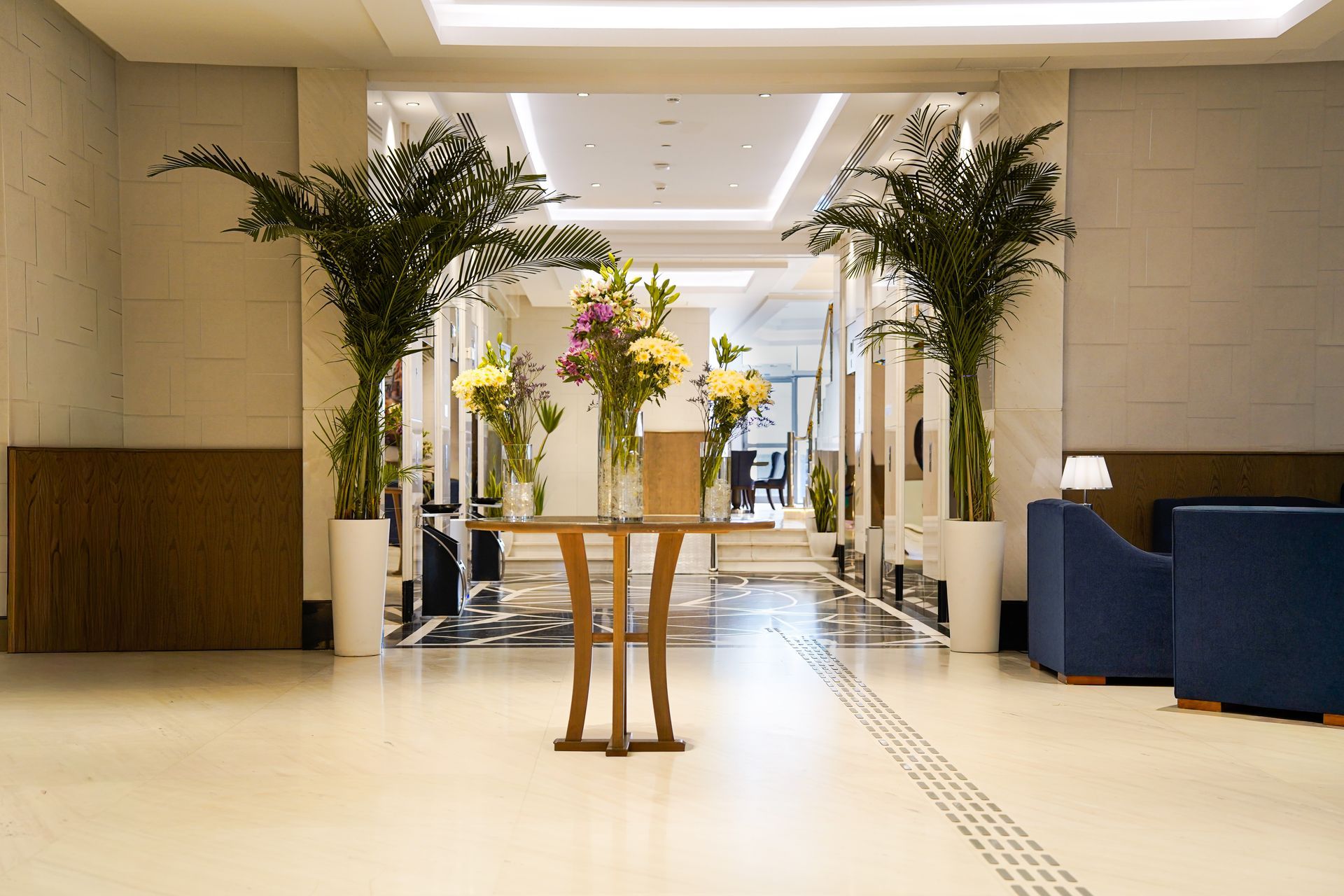 Hotel lobby with flower arrangement on a table, potted palms, and blue seating in a bright hallway