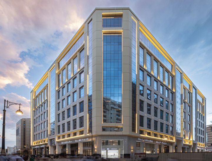 Modern glass-and-stone office building at dusk with lit windows and a city street corner