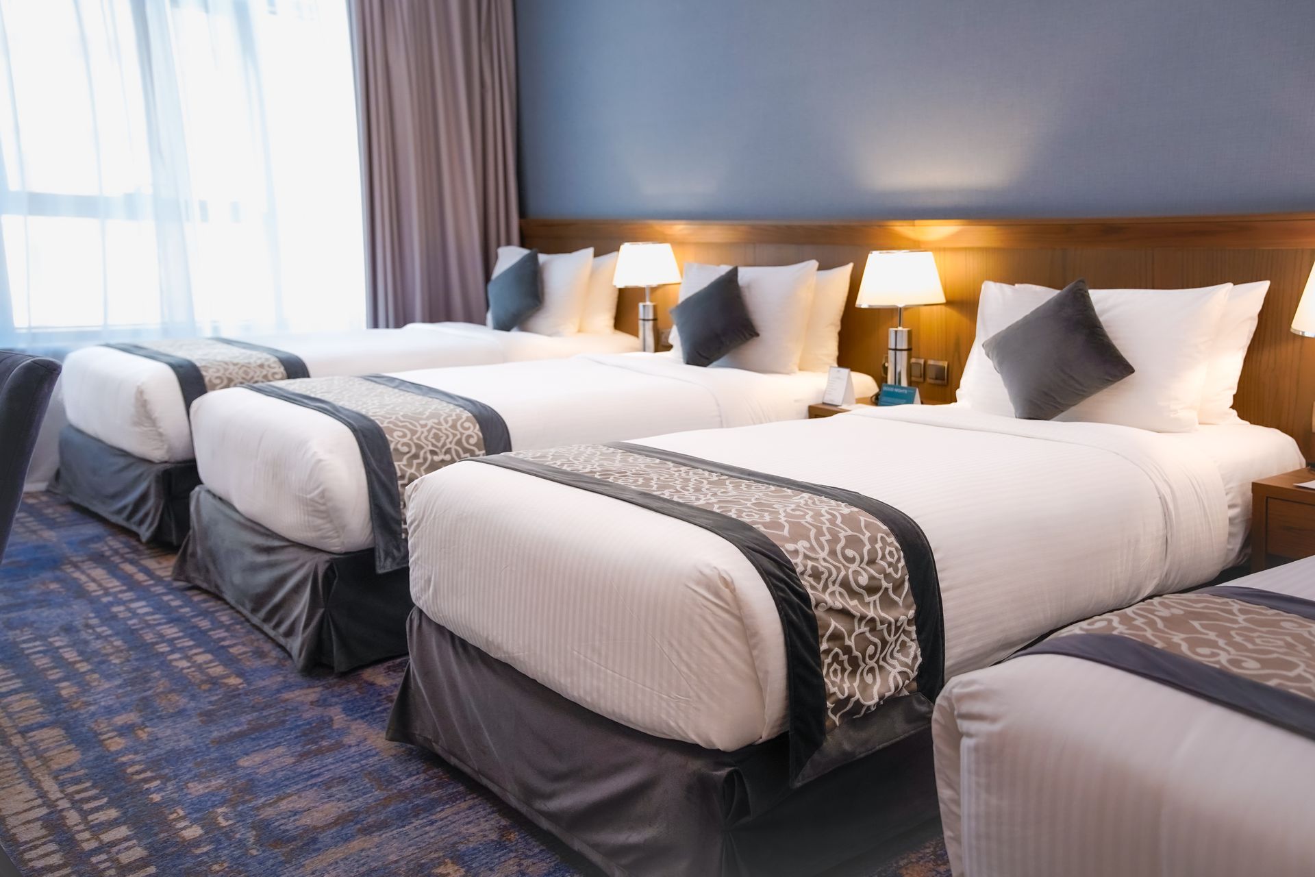 Hotel room with four neatly made beds, white bedding, and blue carpet near a bright window