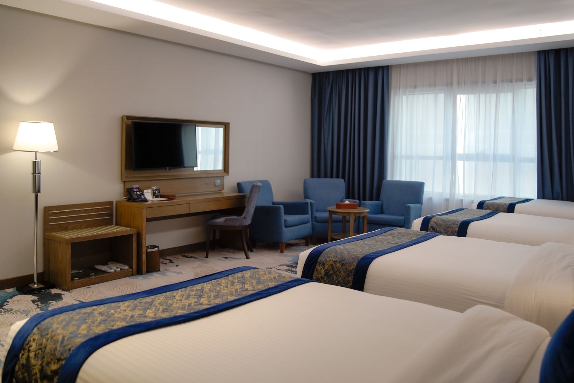 Hotel room with two beds, blue chairs, desk, TV, and large window with curtains