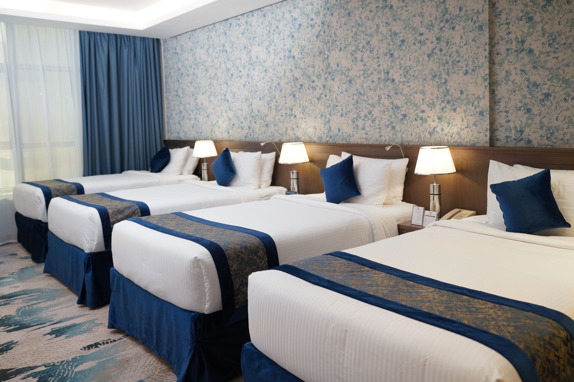 Hotel room with four neatly made twin beds, blue accents, lamps, and floral wallpaper