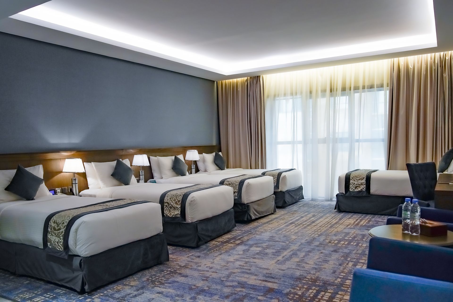 Modern hotel room with four twin beds, blue-gray decor, large window, and curtains
