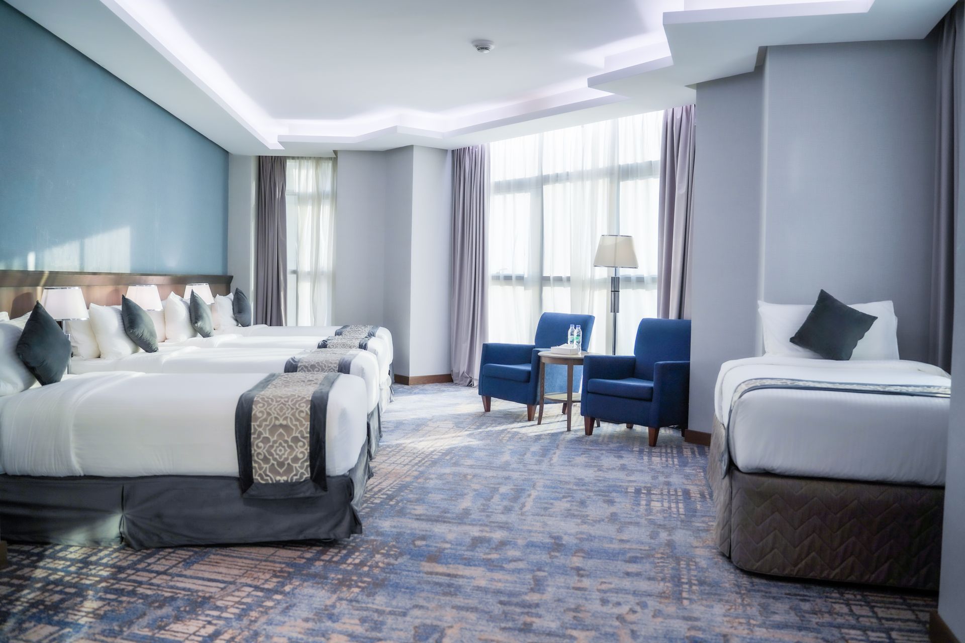 Bright hotel room with two beds, blue chairs by a window, and a patterned carpet.